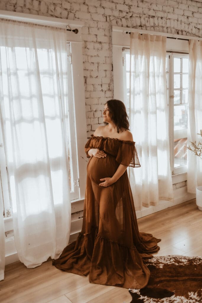 Pregnant mother standing by a window during a maternity photography session near Corona, CA, photographed in a bright natural light studio with soft, neutral tones.