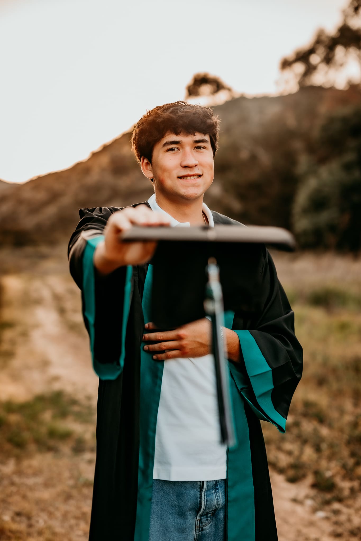 High school senior boy in graduation gown holding cap during golden hour session in Corona California at Skyline Trail