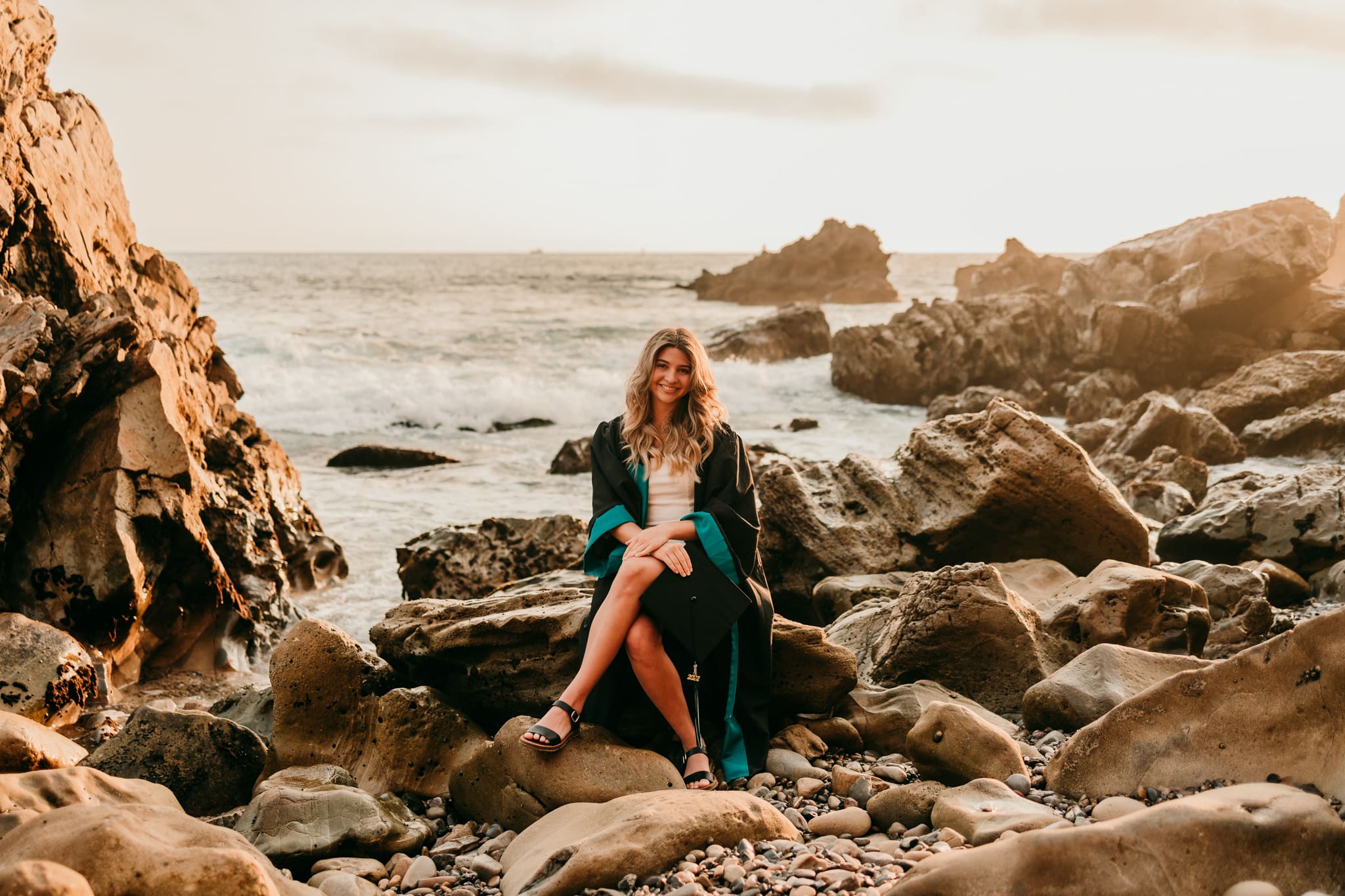 High school senior girl in graduation cap and gown sitting on rocky beach at sunset in Newport Beach California