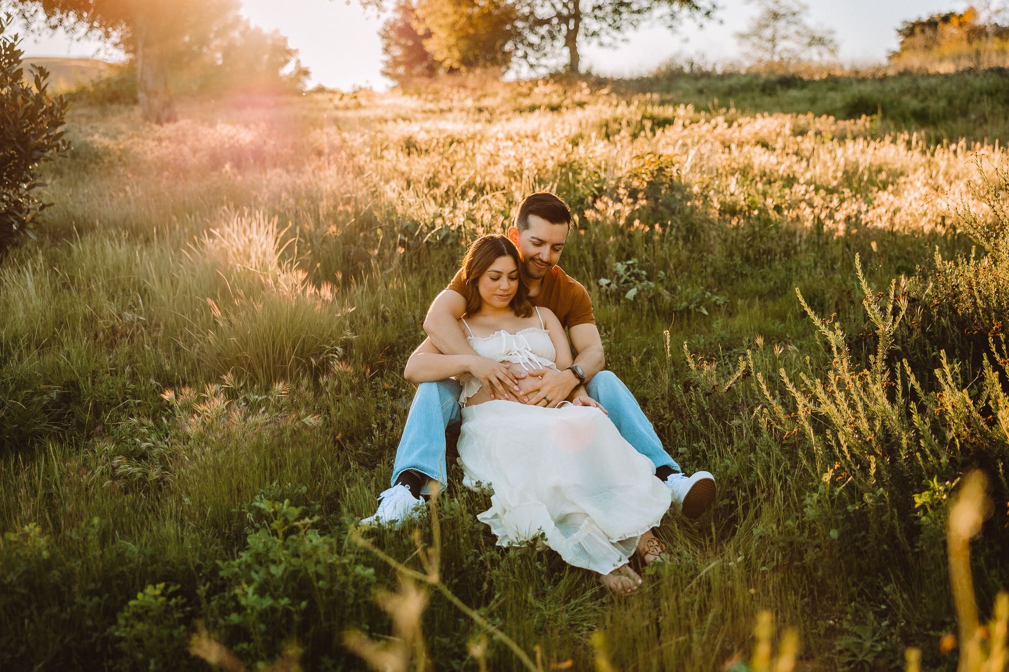 pregnant woman sitting between her husbands legs in the grass during golden hour
