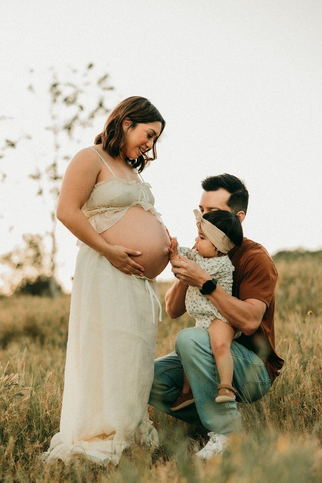 Pregnant mom standing in a field in Chino Hills while her husband and baby kneel next to her baby bump and touch her belly