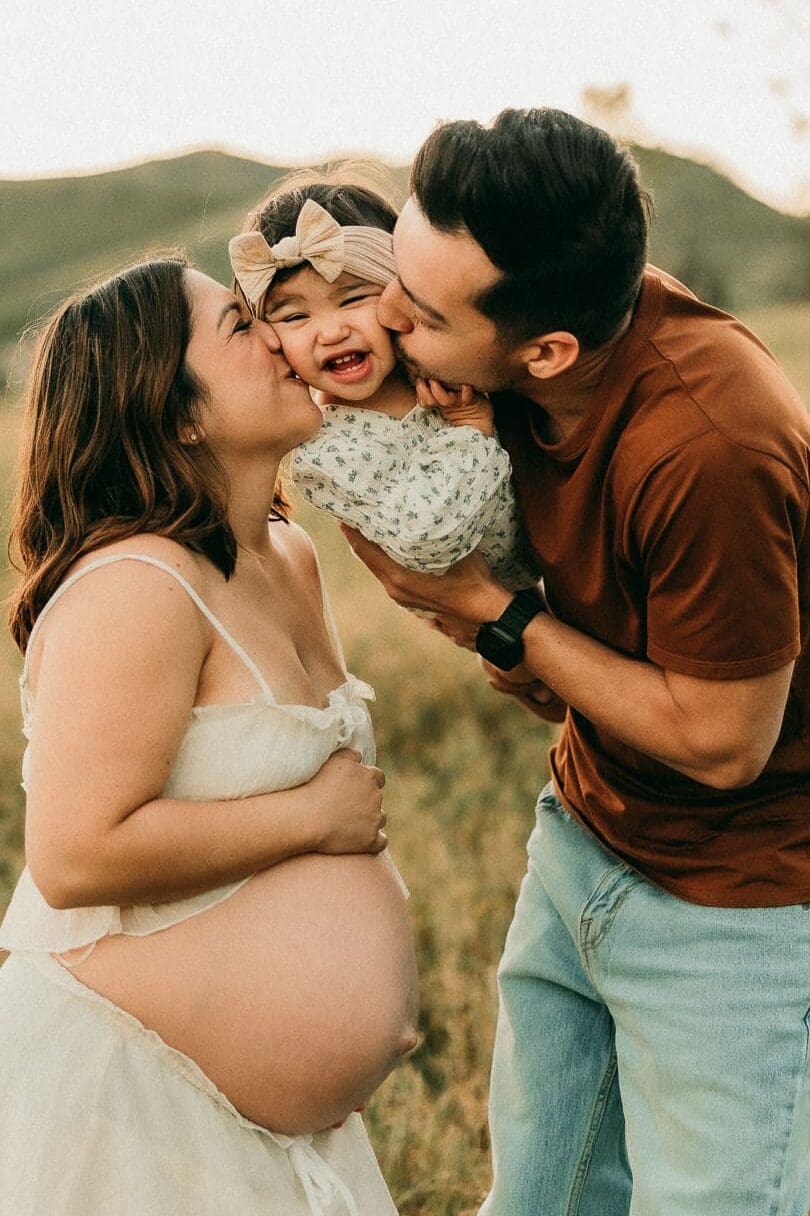 Pregnant mom and husband holding their baby between them and kissing her on the cheek.
