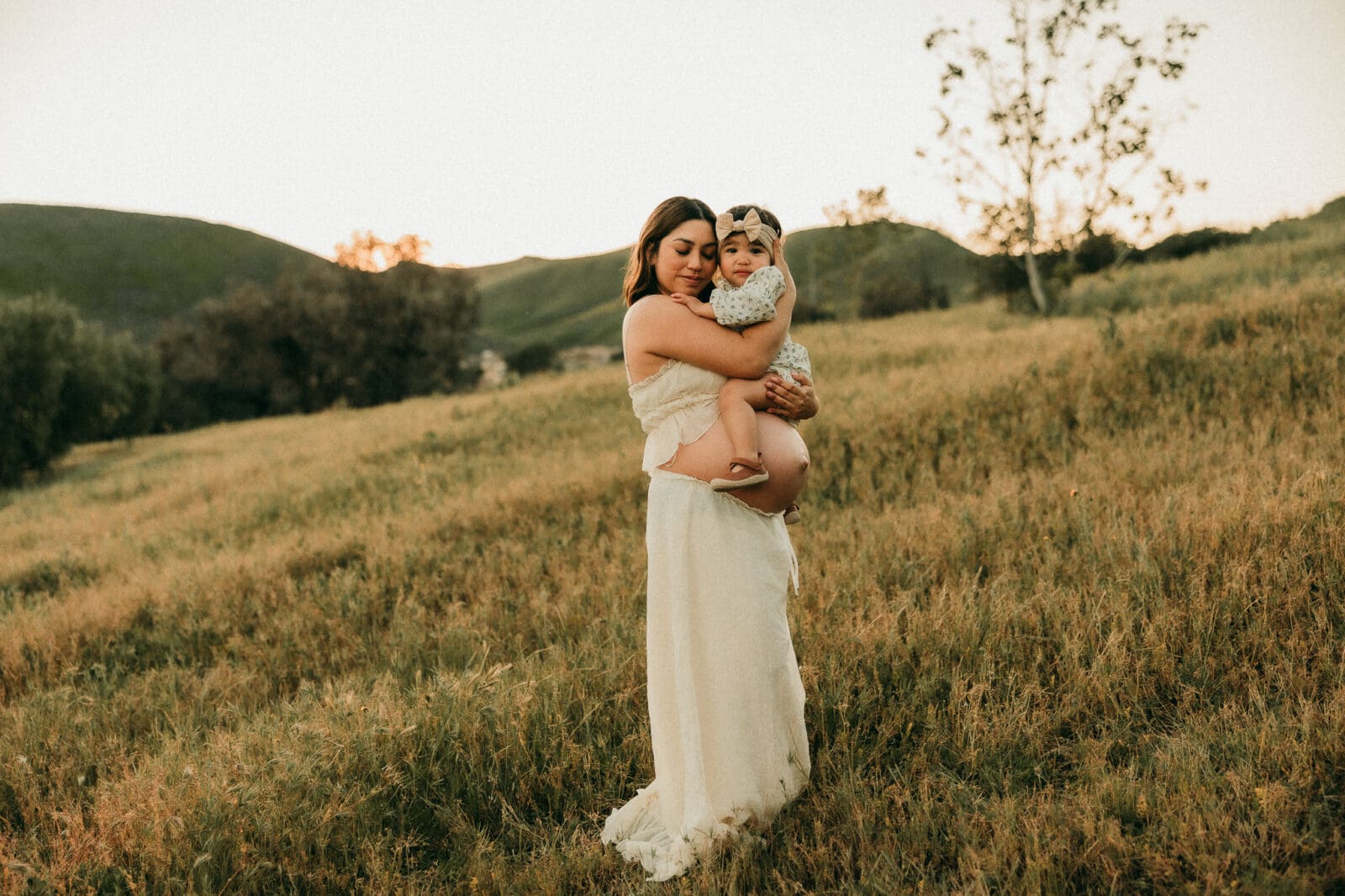 Pregnant mom holding baby in an open field at dusk.