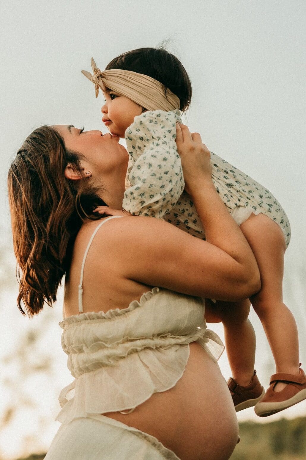 Pregnant mom holding her baby girl above her and giving her a kiss on her chin