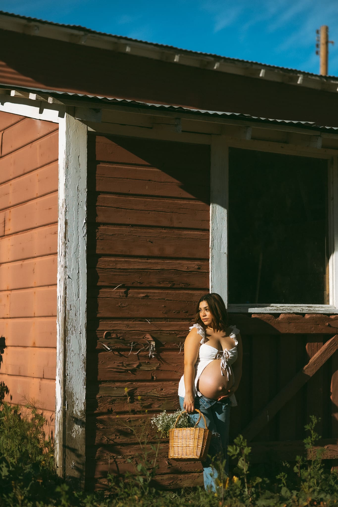 Pregnant woman holding a basket of flowers standing up against a barn and looking into the sunlight