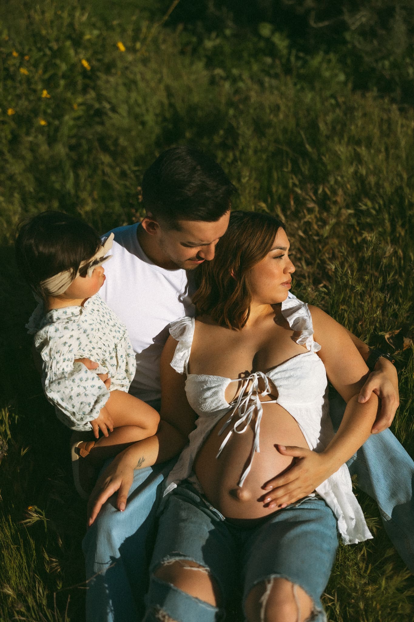 Pregnant woman laying on her husbands lap with their baby in a grassy field